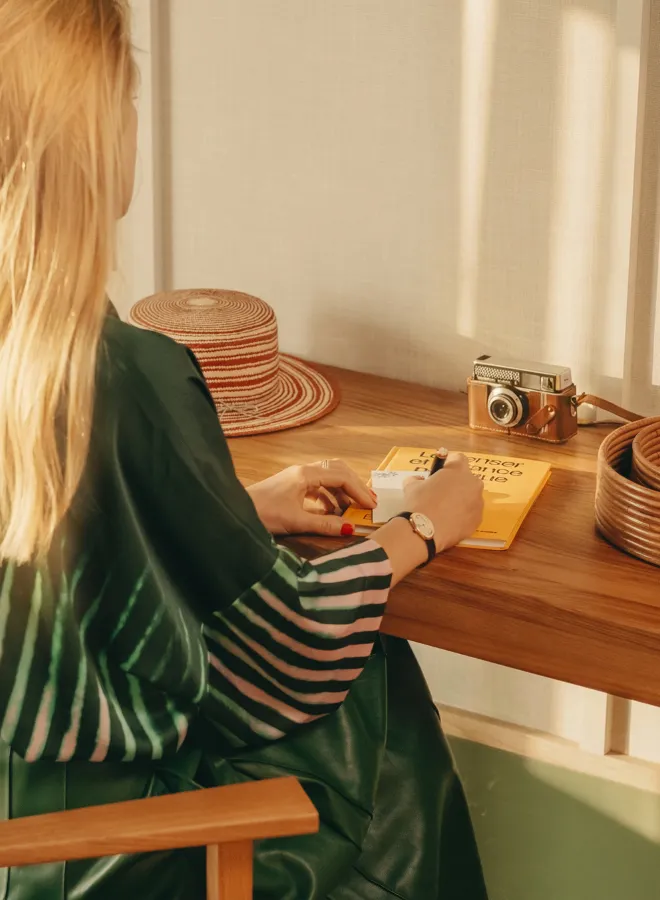 Femme aux cheveux blonds assise à une table en bois, écrivant sur un bloc-notes avec un chapeau en paille et un appareil photo vintage posés sur la table.
