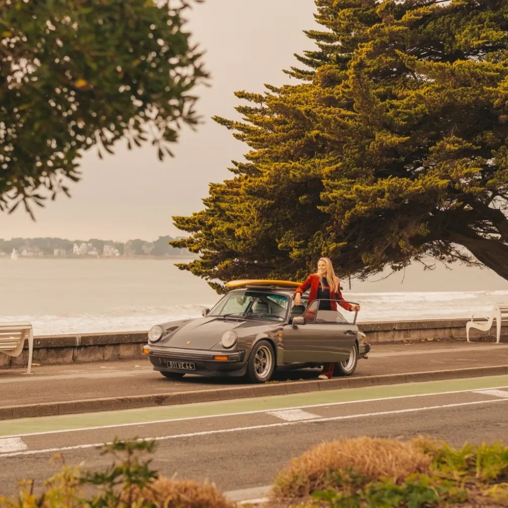 Femme blonde souriante debout à côté d'une voiture de sport grise classique garée au bord de la mer avec une planche de surf sur le toit.