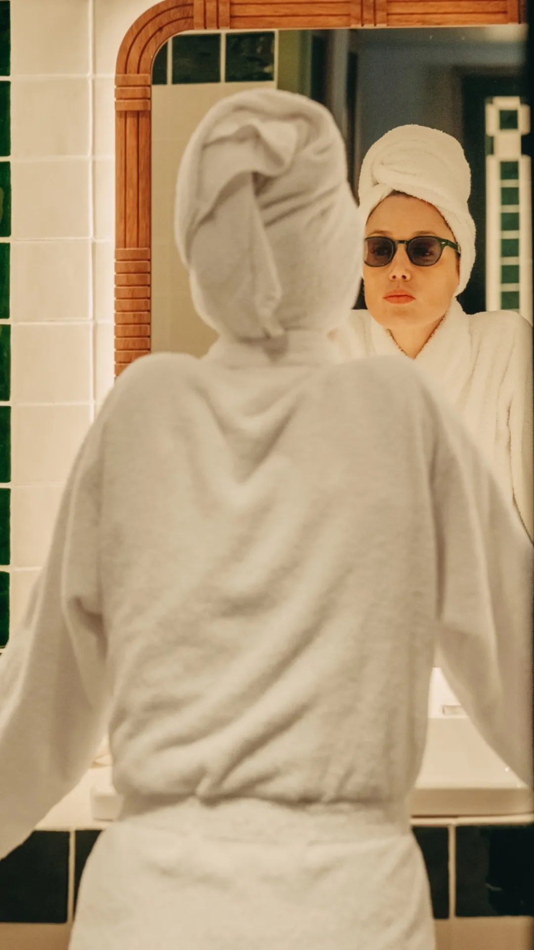 Femme portant un peignoir et une serviette sur la tête, regardant son reflet dans un miroir de salle de bain en portant des lunettes de soleil.