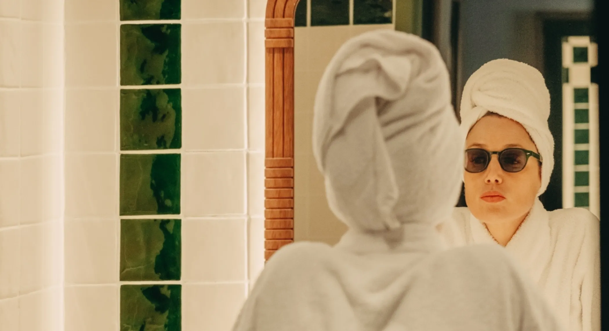 Femme avec un peignoir blanc et une serviette sur la tête regarde dans un miroir, portant des lunettes de soleil dans une salle de bain avec des carreaux verts et blancs.