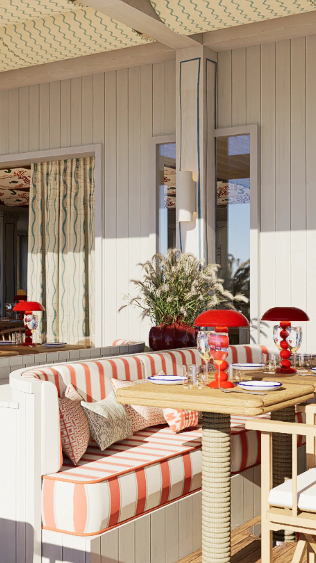 Coin repas lumineux avec banquette rayée orange et blanche, coussins décoratifs, table en bois et lampes rouges sur une terrasse ensoleillée.