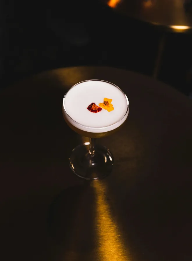 Cocktail with white foam and two red and yellow edible flowers on a dark table.