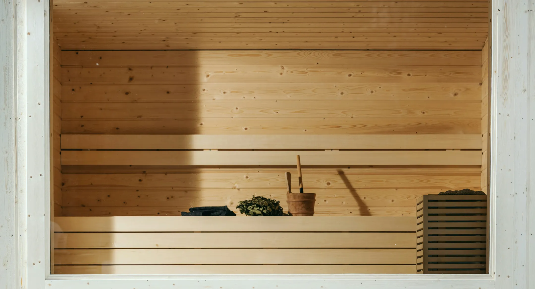 Intérieur d'un sauna en bois clair avec un seau, une louche et un bouquet de feuilles sur le banc.