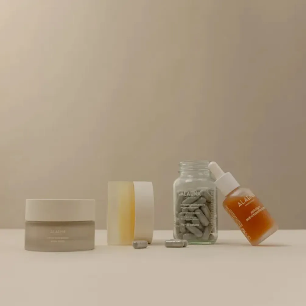 Quatre produits de soins sur une surface beige: un pot de crème, un savon transparent avec un couvercle blanc, un flacon en verre avec des gélules grises, et une bouteille avec un compte-gouttes à moitié inclinée.