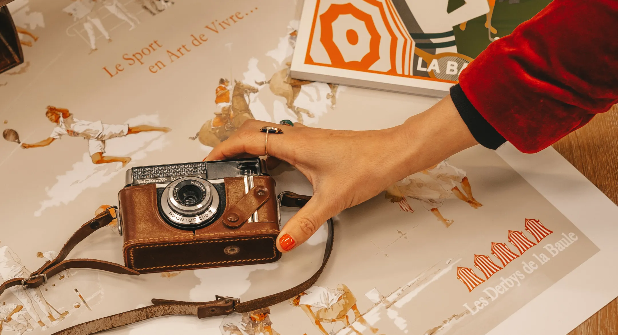 Main tenant un appareil photo vintage avec étui en cuir posé sur une affiche illustrant des sports et le texte « Les Derbys de la Baule ».