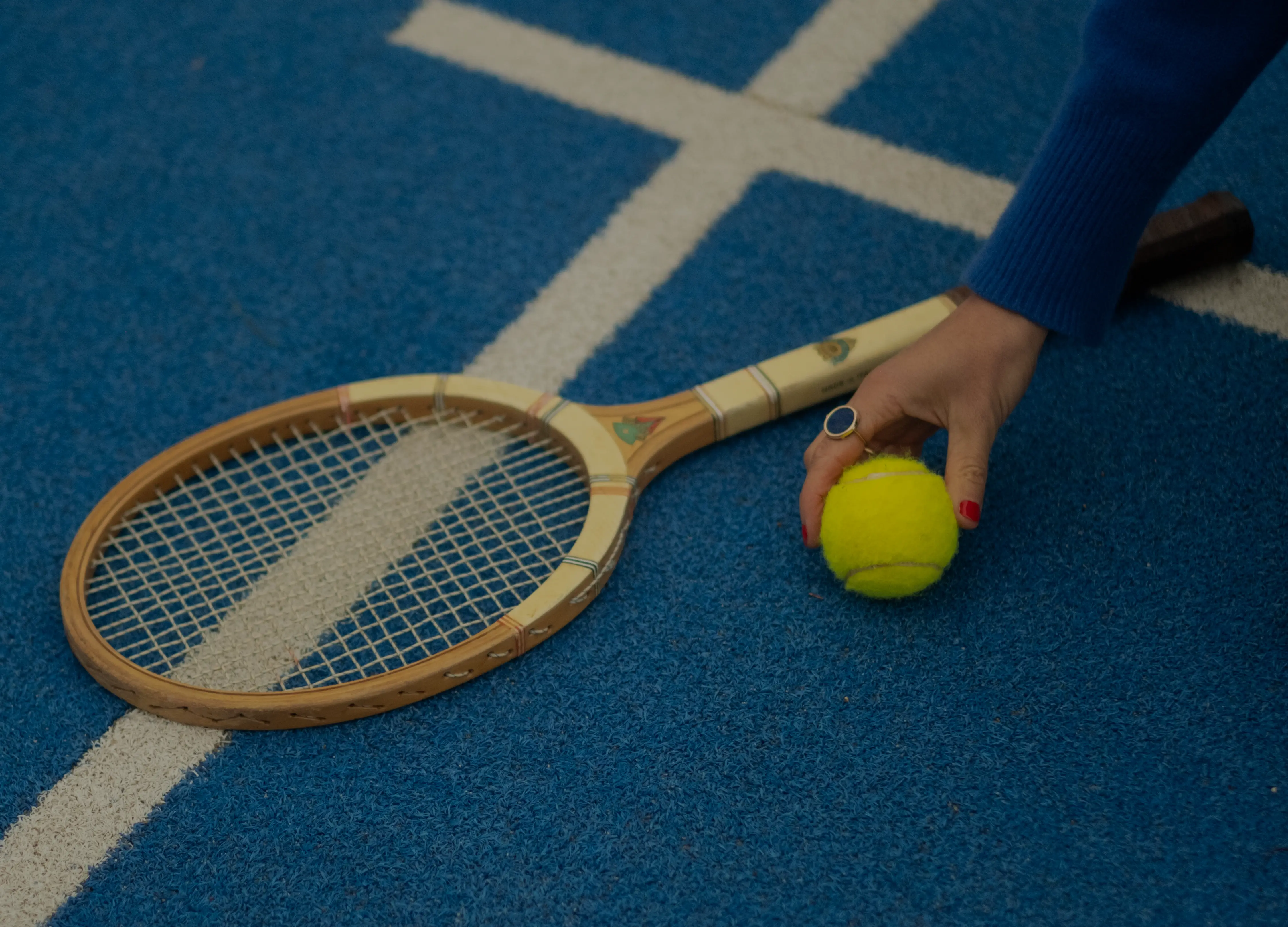 Main tenant une balle de tennis jaune près d'une raquette de tennis en bois sur un court bleu avec des lignes blanches.