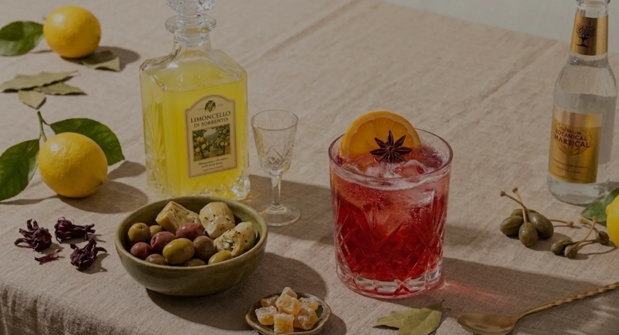 Cocktail rouge glacé dans un verre taillé avec une tranche d'orange et une étoile d'anis, accompagné d'une bouteille de limoncello, un bol d'olives et artichauts, des citrons, des câpres, et une petite assiette de morceaux d'orange confite.