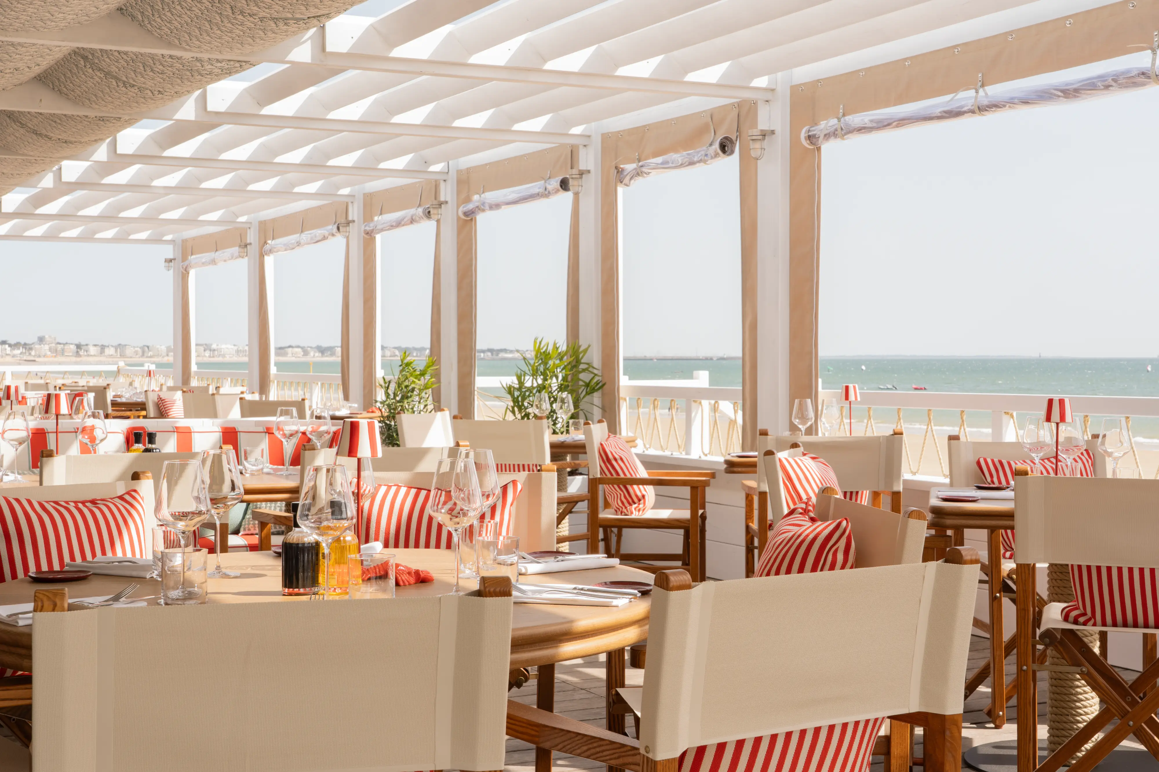 Terrasse de restaurant en bord de mer avec tables en bois, chaises à rayures rouges et blanches, et vue sur l'océan.