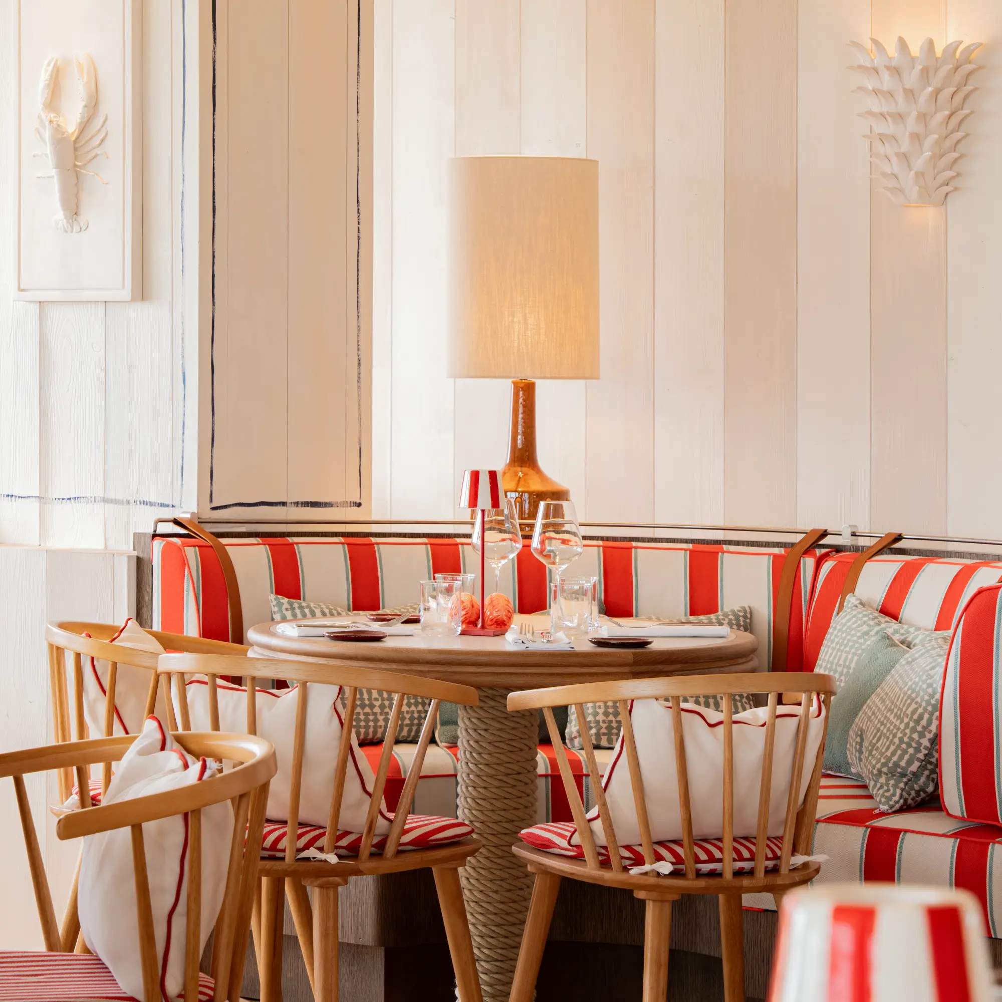 Coin repas chaleureux avec une table ronde en bois, chaises en bois clair avec coussins blancs et rouges, banquette rayée rouge et beige, lampe à abat-jour beige et décorations murales blanches.