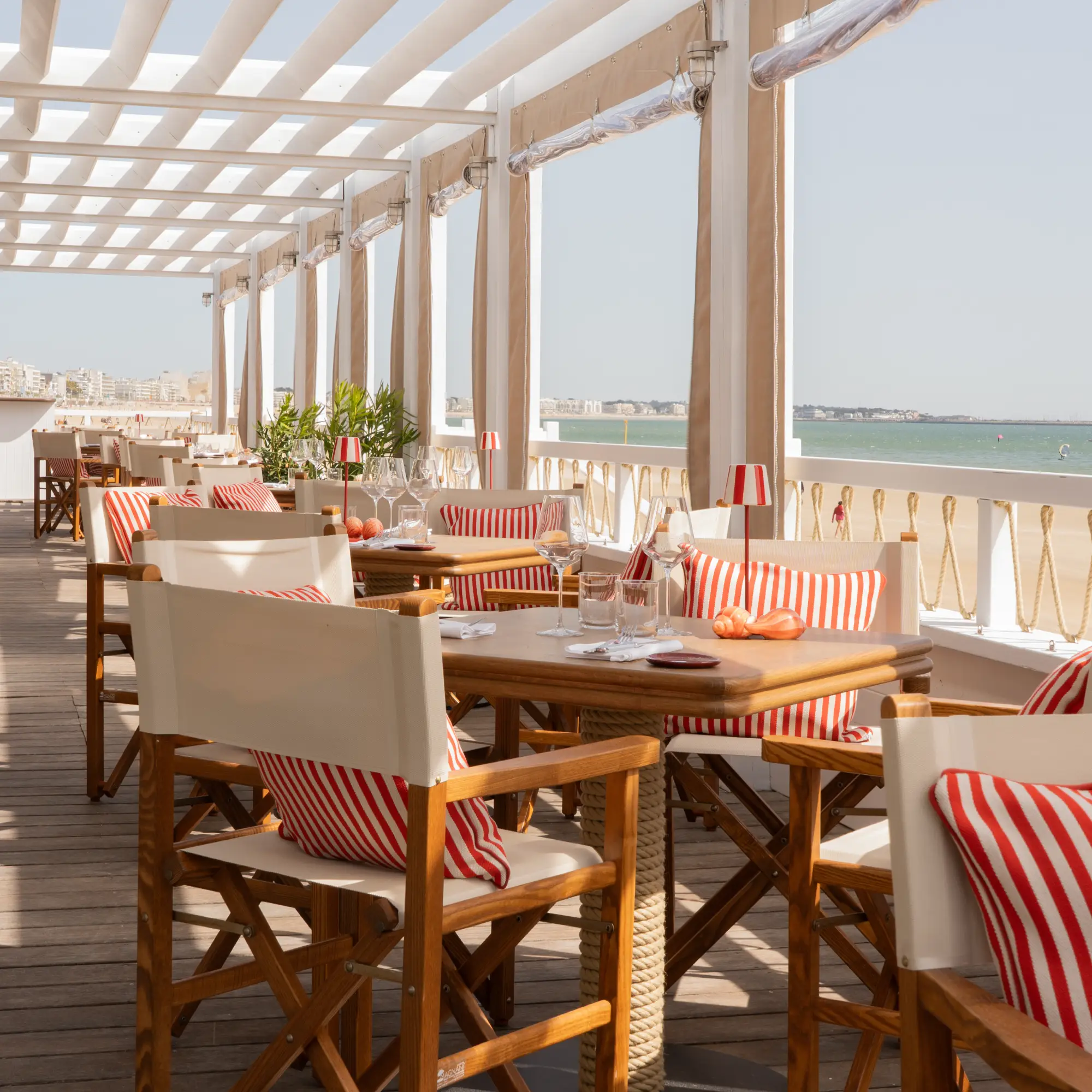 Terrasse d'un restaurant en bois avec des chaises en toile blanche et coussins rayés rouges, vue sur la plage et la mer au loin.