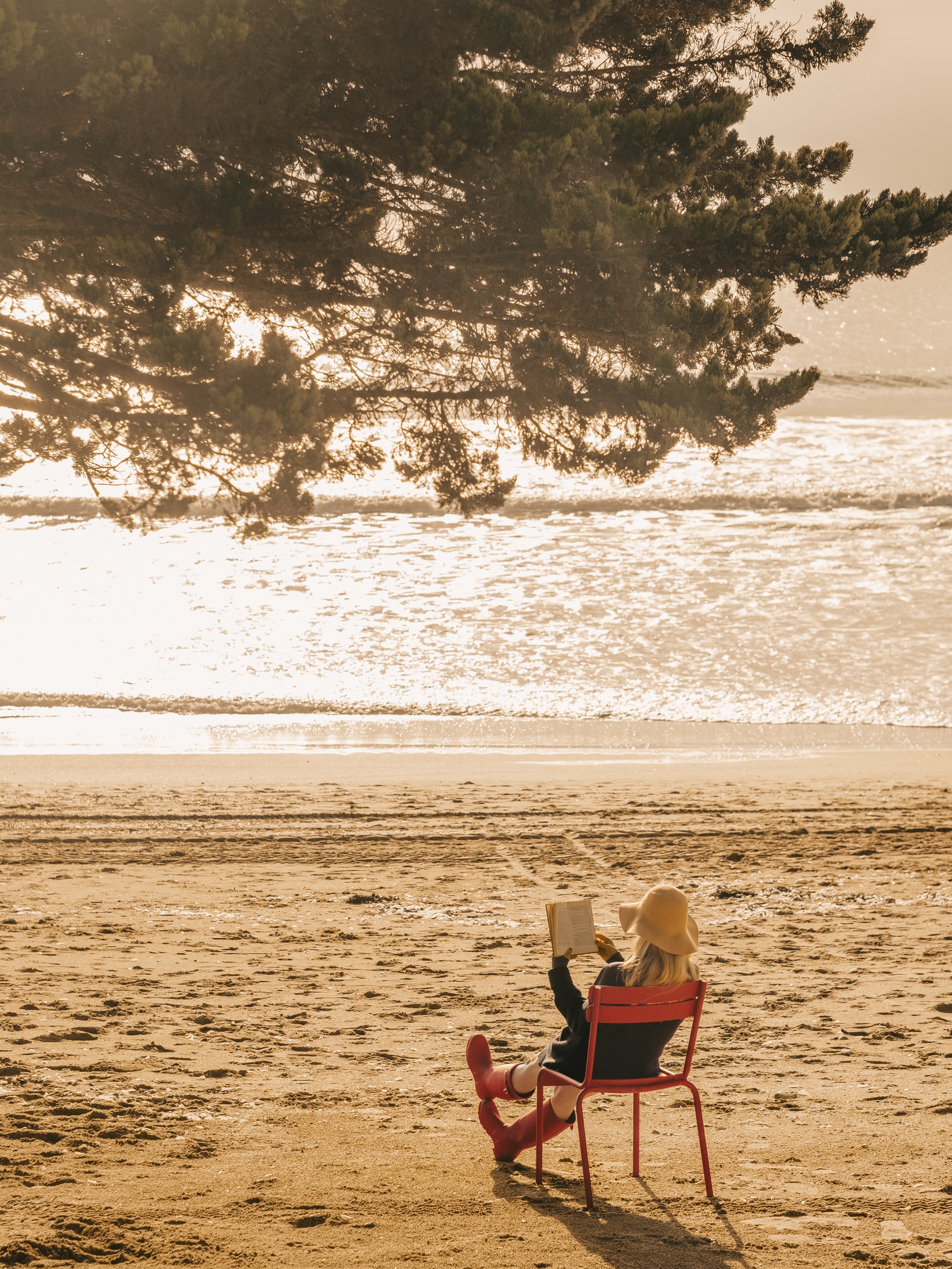 Personne assise sur une chaise rouge sur la plage, lisant un livre sous un arbre avec vue sur la mer bauloise scintillante.