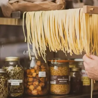 Des pâtes fraîches longues suspendues sur une étagère en bois au-dessus de bocaux en verre remplis d'herbes, champignons et condiments dans une cuisine rustique.