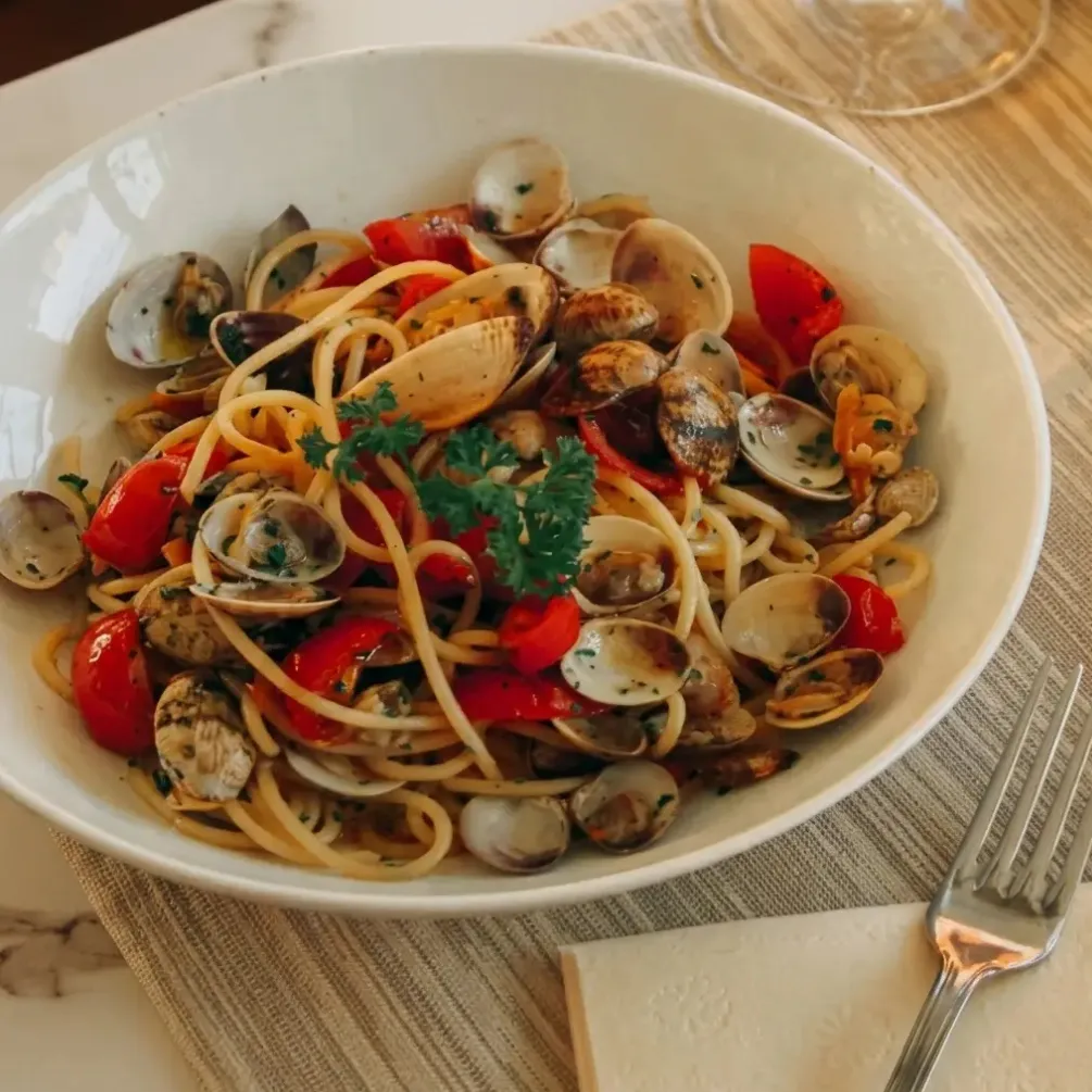 Assiette de spaghetti aux palourdes, tomates cerises et persil sur une table avec une fourchette et une serviette.