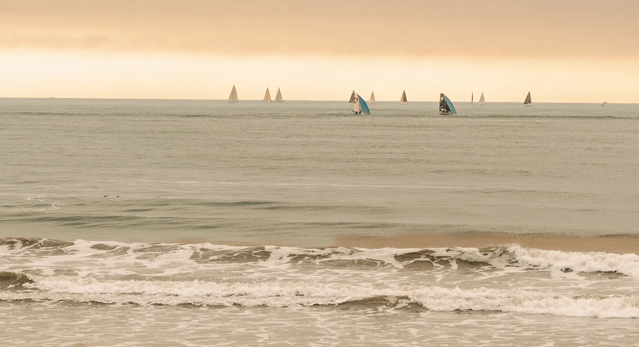 Calm sea with several white and blue sailboats under an orange sky at sunset.