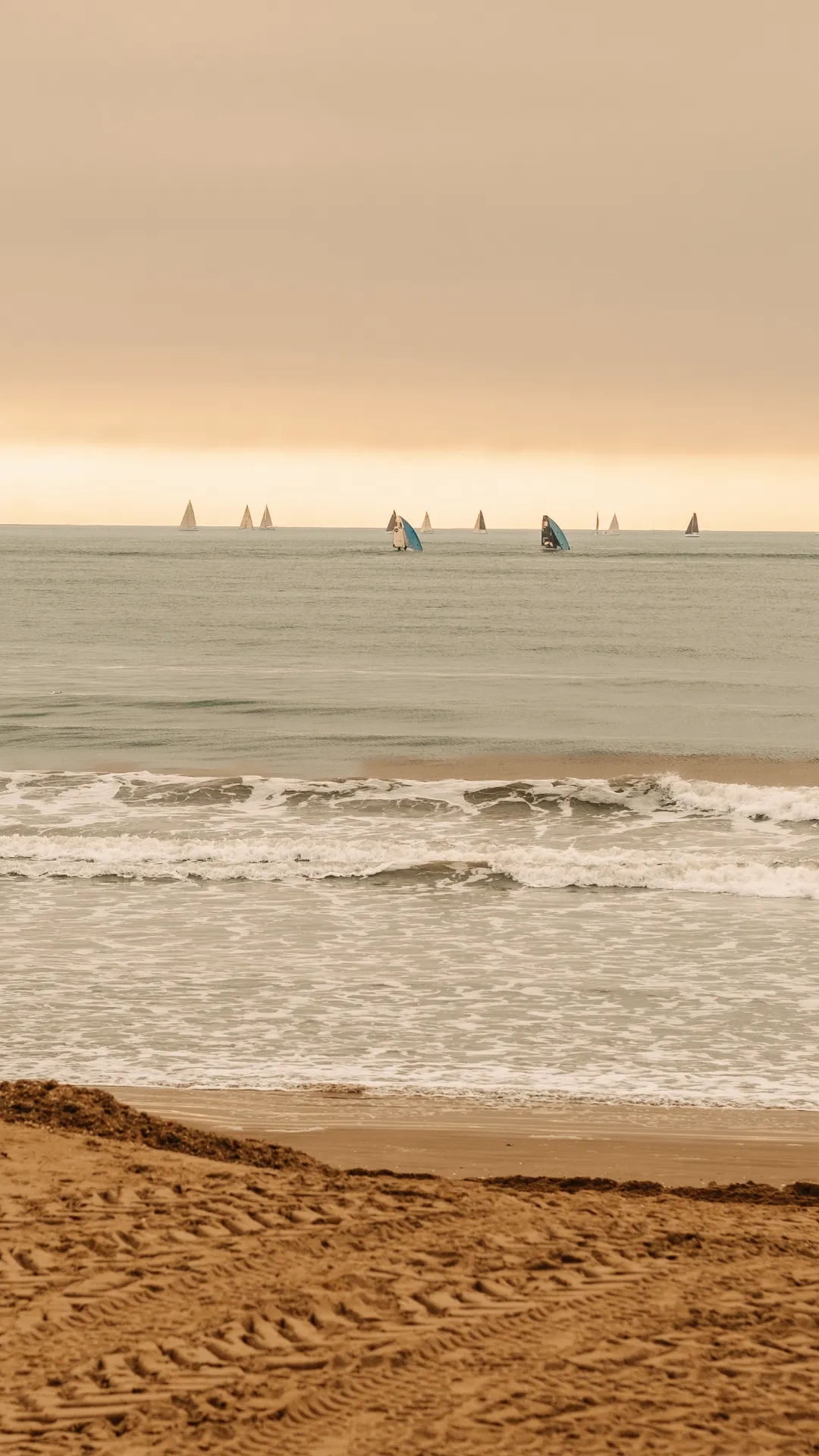 Sandy beach with several sailboats on the calm water under a cloudy sky at sunset.