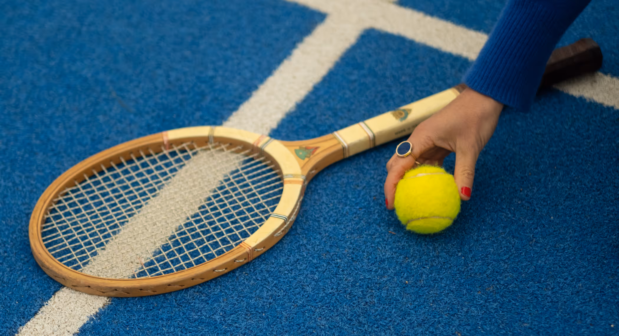Main tenant une balle de tennis jaune à côté d'une ancienne raquette de tennis en bois sur terrain bleu.