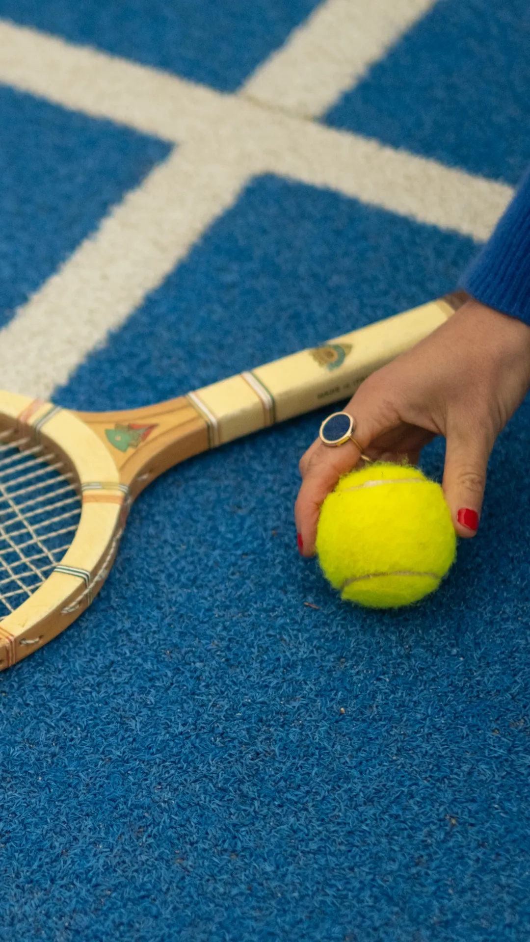 Main tenant une balle de tennis jaune, raquette de tennis sur sol bleu avec lignes blanches.