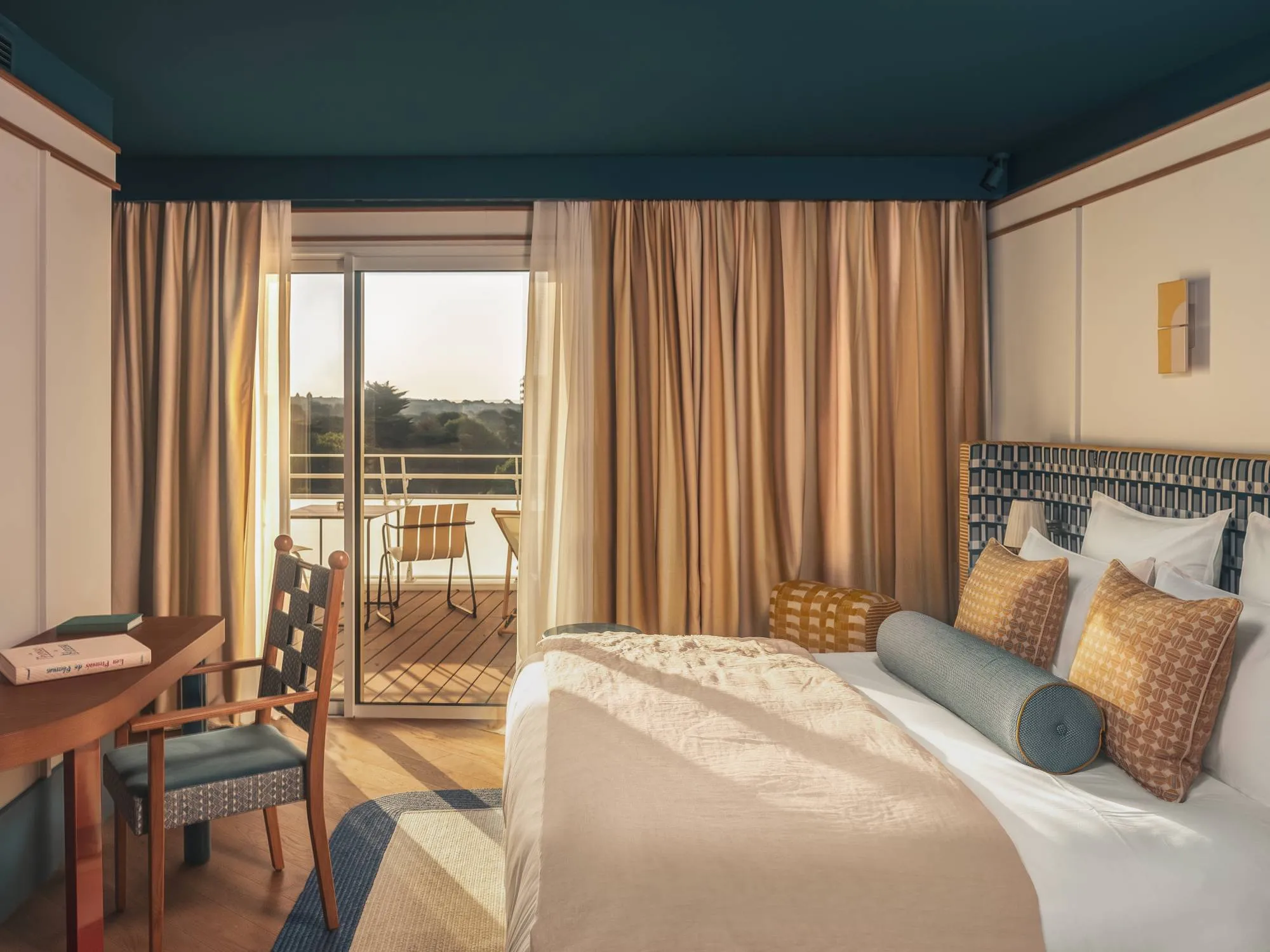 Room with bed, table and chair near a patio door leading to a sunny balcony.