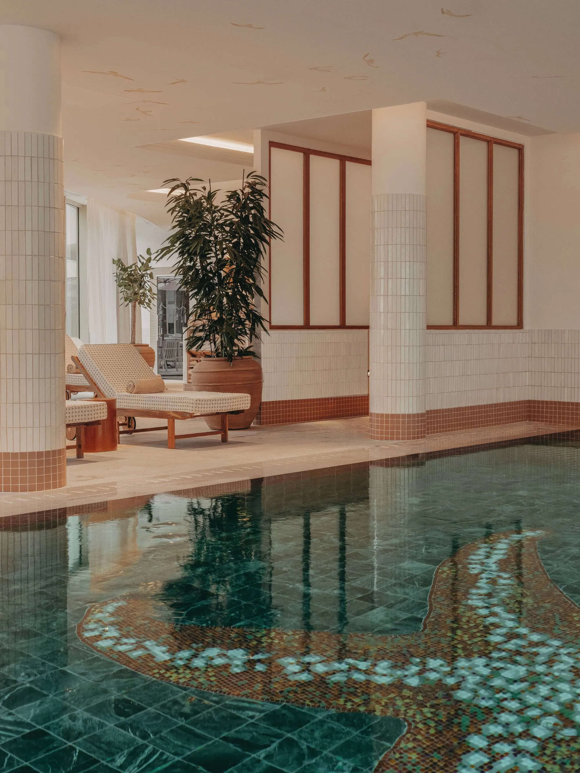 Indoor pool with green tiling and beige lounge chair next to large potted plants.