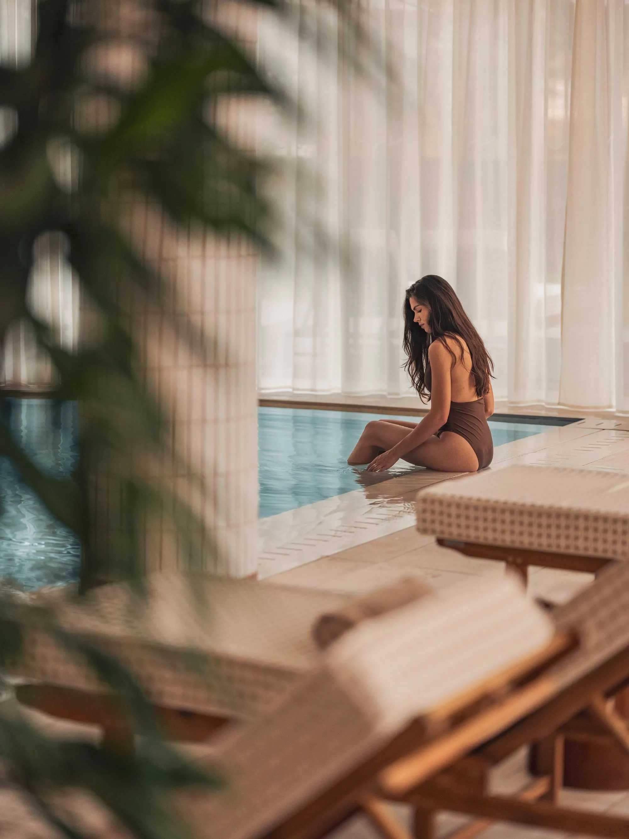 Woman in brown swimsuit sitting by an indoor pool with fuzzy lounge chairs.