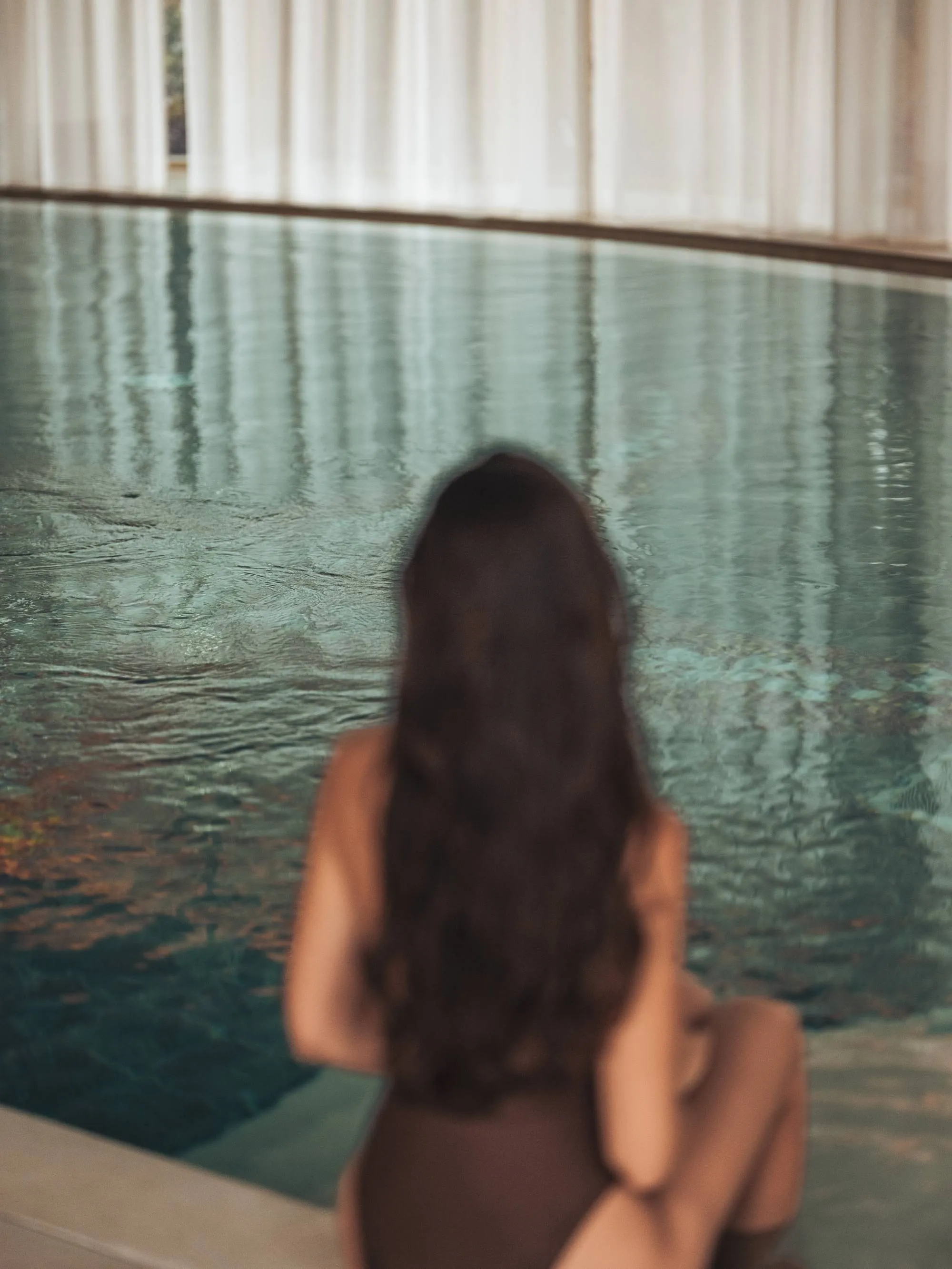 Woman sitting by an indoor pool with long black hair seen from the back.