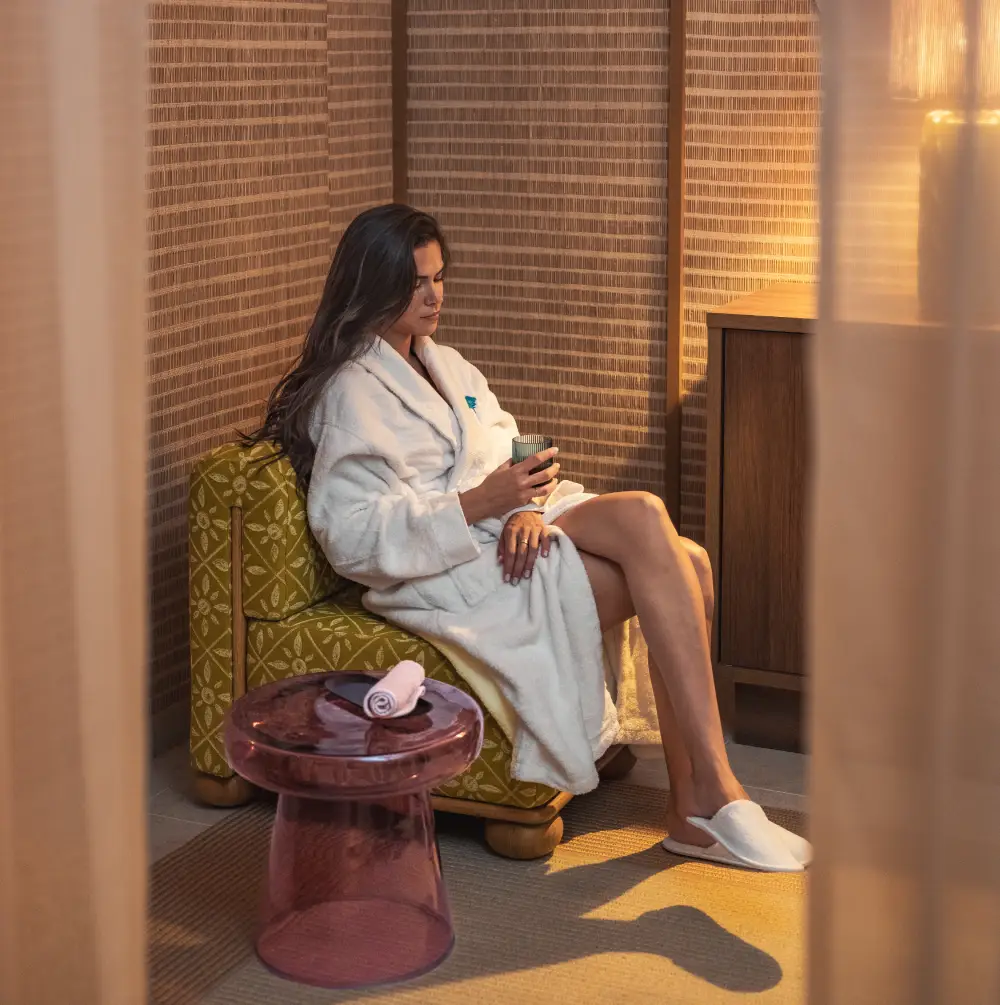 Woman in a white bathrobe sitting on a green armchair, holding a cup in a warmly decorated living room.