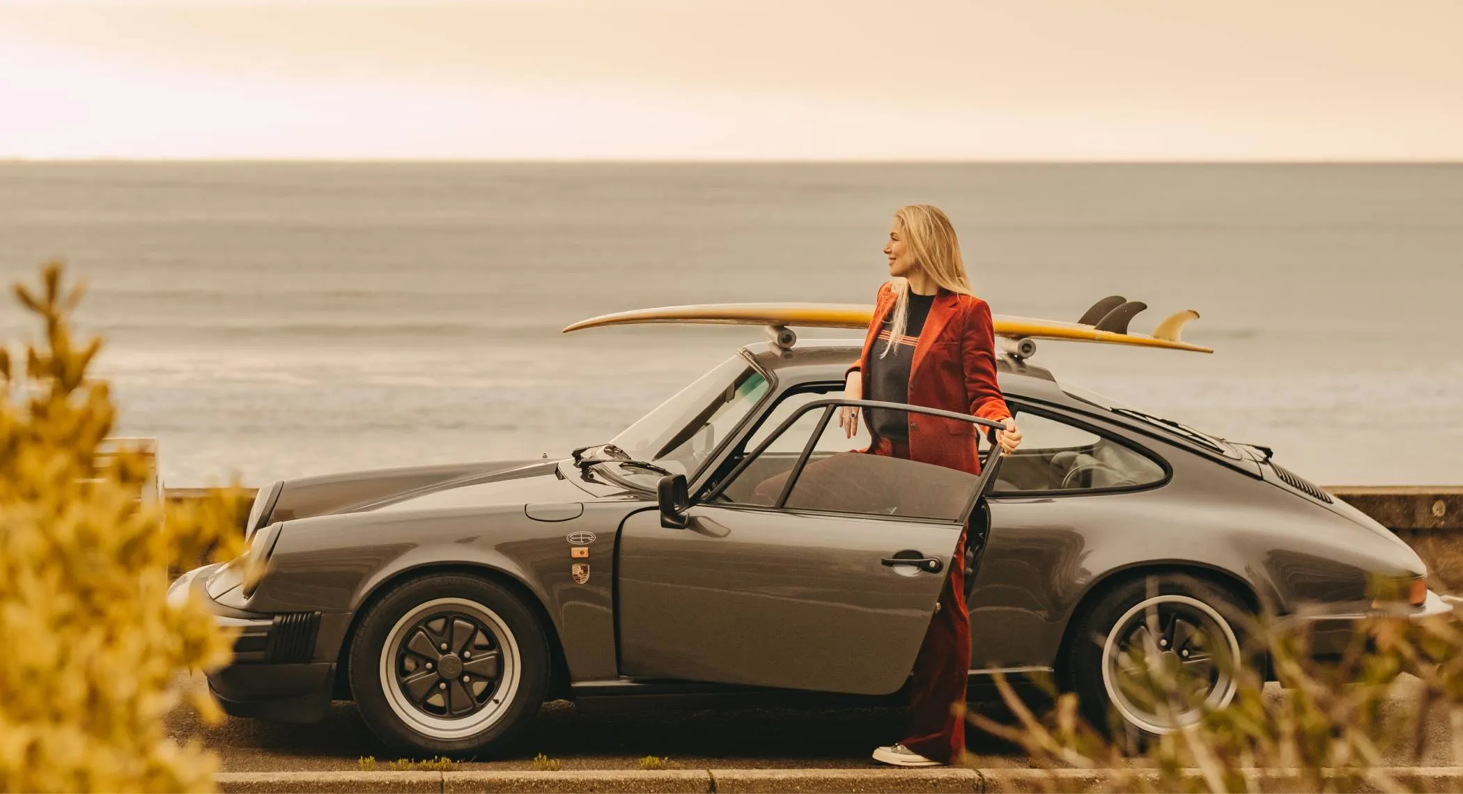 Femme en costume rouge debout près d'une voiture grise avec une planche de surf jaune sur le toit.