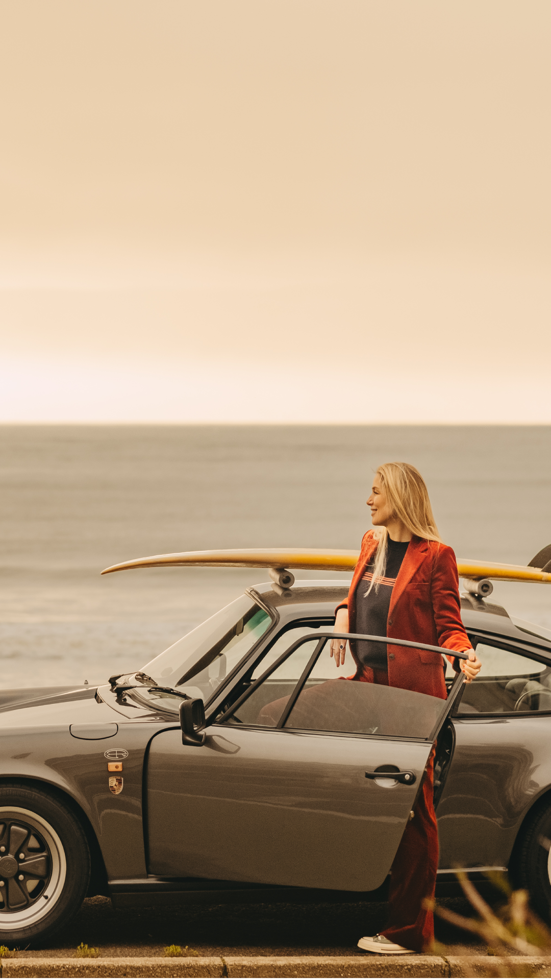 Femme en tailleur rouge ouvrant la portière d'une voiture grise près de la mer avec une planche de surf.