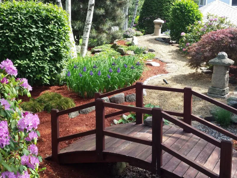 Wooden arched garden bridge over gravel path surrounded by green shrubs, purple irises, and flowering plants in a landscaped garden.