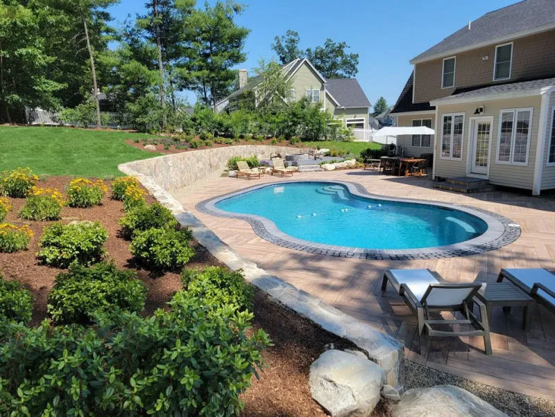 Backyard with a kidney-shaped swimming pool surrounded by a brick patio, lounge chairs, and landscaped bushes with two houses in the background.