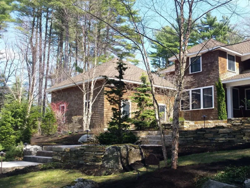 Two-story wooden house surrounded by tall pine trees and landscaped stone retaining walls in a forested area.