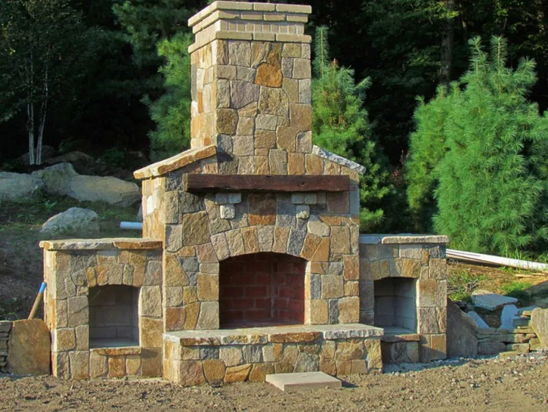 Outdoor stone fireplace with a central chimney and two smaller arches on either side, set in a natural wooded area.