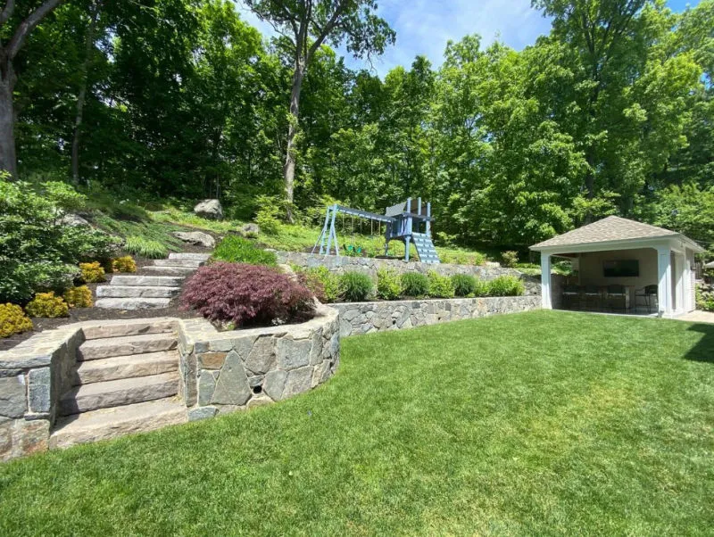 Backyard with green lawn, stone retaining walls, wooden stairs, a children's playset, and a small covered patio area with seating.