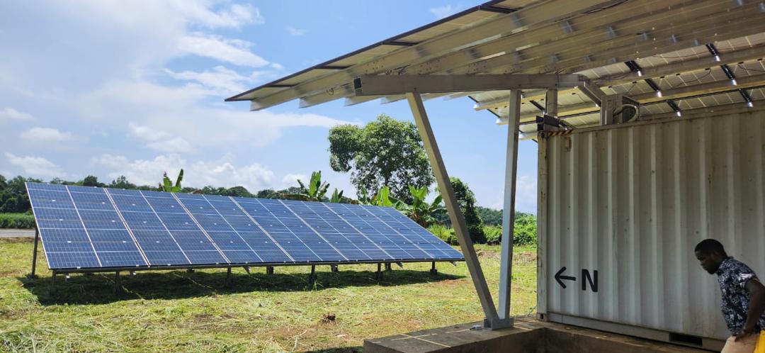 Solar panels mounted on a metal frame in a grassy field next to a white shipping container with a man standing nearby under a solar panel-covered roof.