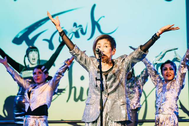 Kazuko Hokhi, a short Japanese woman with an asymmetrical bob haricut, stands on stage in front of a microphone. She wears a silver jacket and her hands are raised up in the air.