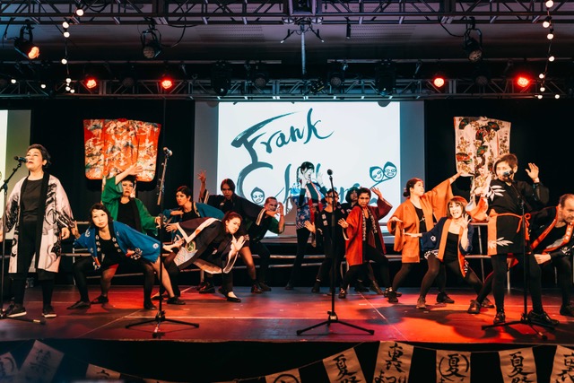 A group of people singing and dancing together on stage. In the background can be seen a banner reading 'Frank Chickens'