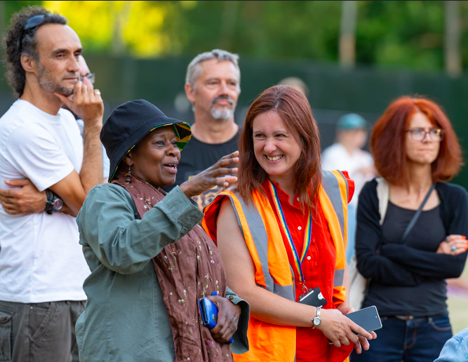 A woman in an orange high-vis vest that reads 'volunteer' smiles as she talks to another woman, who gestures excitedly at something off-camera.