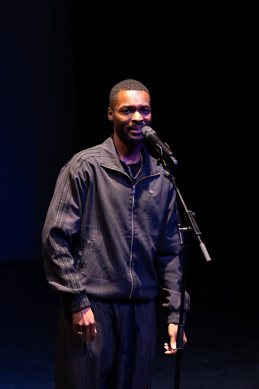 Haringey based poet, Kieron Rennie. He is a young Black man with short hair, wearing a pinstriped-black tracksuit. He is performing on stage, speaking into a microphone. There are black curtains behind him, and he is under a spotlight. 
