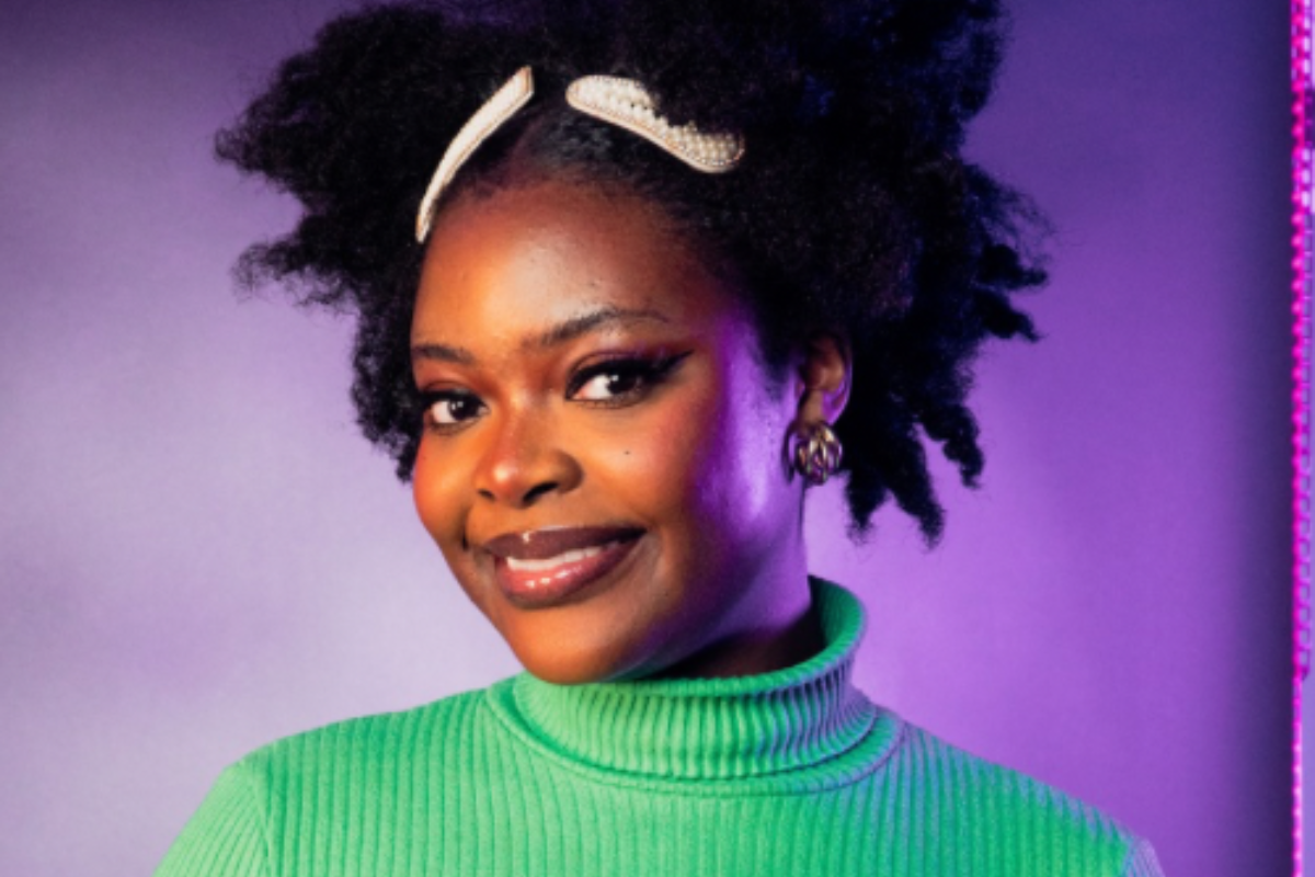 A professional headshot of a Black woman wearing Afro hair decorated with pearly clips. She has impeccable eyeliner and wears an emerald green sweater