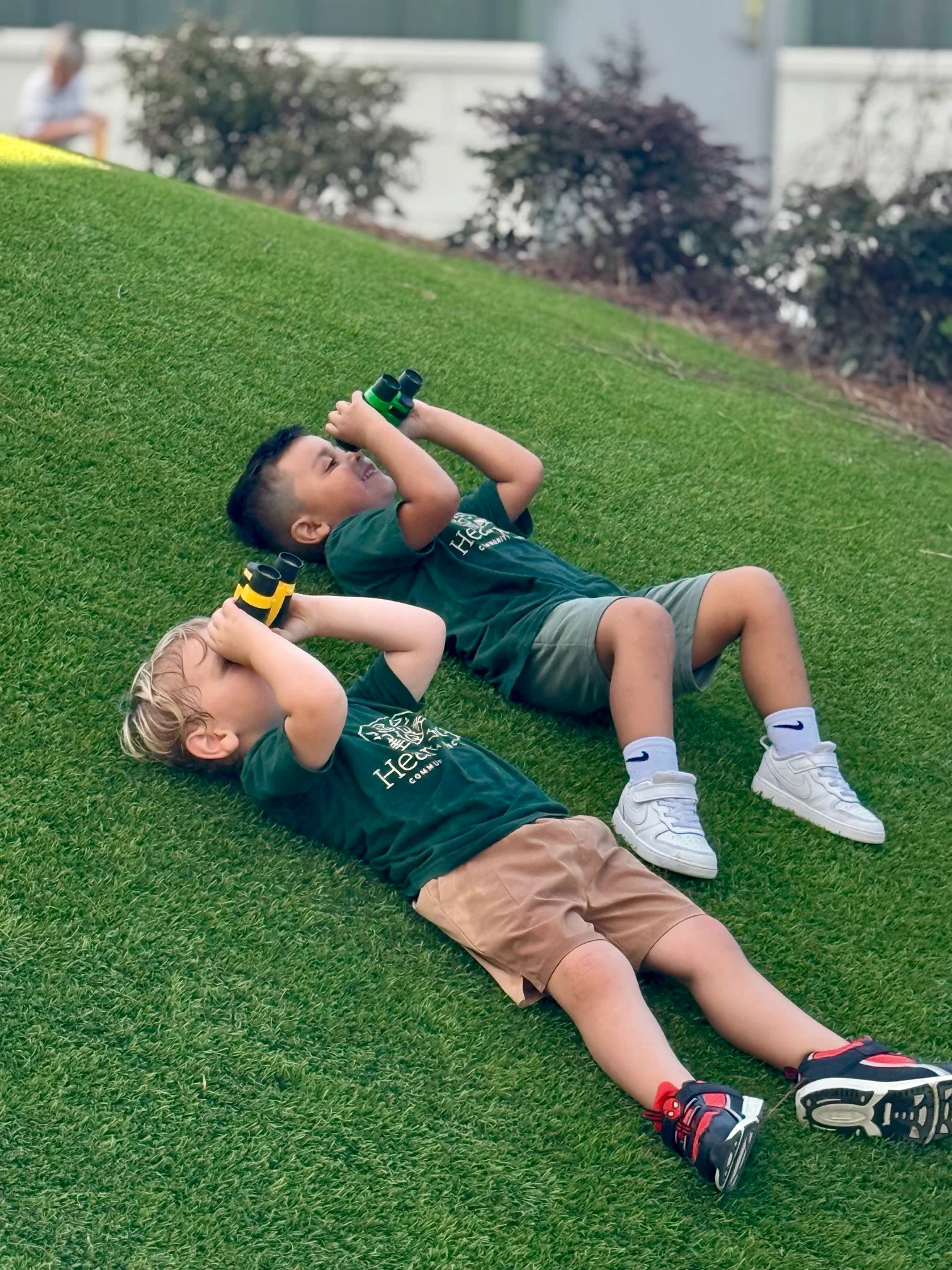 Two young boys lying on grass looking through binoculars, wearing green shirts and shorts.