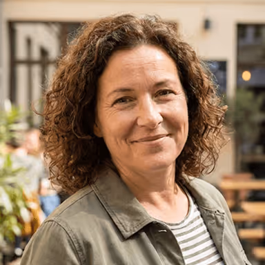 Frau mit lockigen Haaren lächelt im Freien, trägt eine gestreifte Bluse und olivgrüne Jacke.