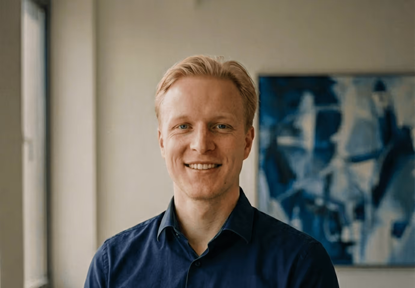 Lächelnder Mann mit blonden Haaren und blauem Hemd vor abstraktem Gemälde in modernem, lichtdurchflutetem Raum.