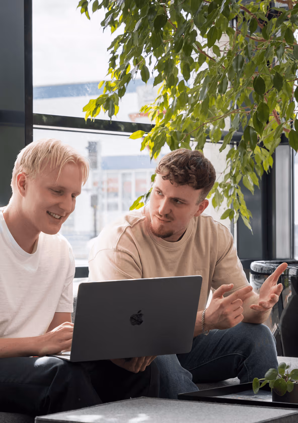 Zwei Männer arbeiten lächelnd an einem Laptop in einem modernen Büro mit großen Fenstern und Pflanzen.