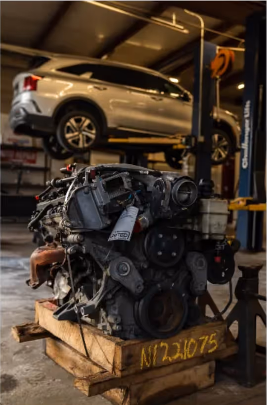 Car engine placed on a wooden pallet inside a garage with a silver SUV elevated on a hydraulic lift in the background.