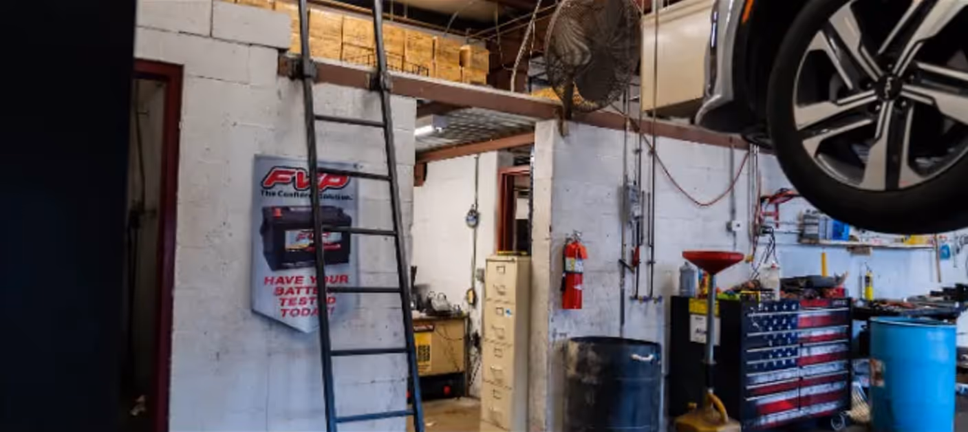 Interior of an auto repair shop with a black ladder against a wall, a filing cabinet, a fire extinguisher, a large wall fan, an American flag tool chest, and a car lifted overhead.