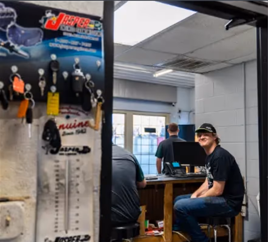 Two men seated at a workbench with computer monitors in a workshop, and a key holder with multiple keys hanging on the left side.