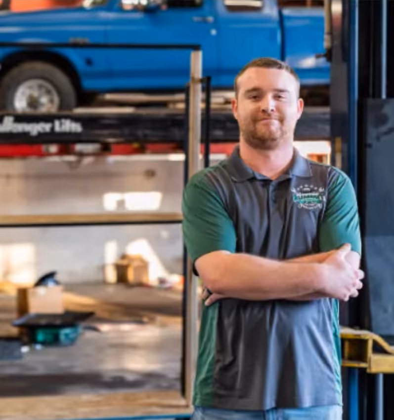 Mechanic standing with arms crossed in a workshop, with a blue pickup truck raised on a lift behind him.