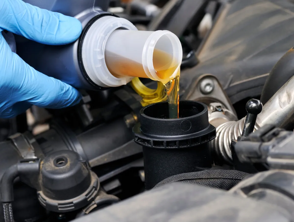 Hand wearing blue glove pouring golden engine oil into a black engine oil filler cap in a car engine.