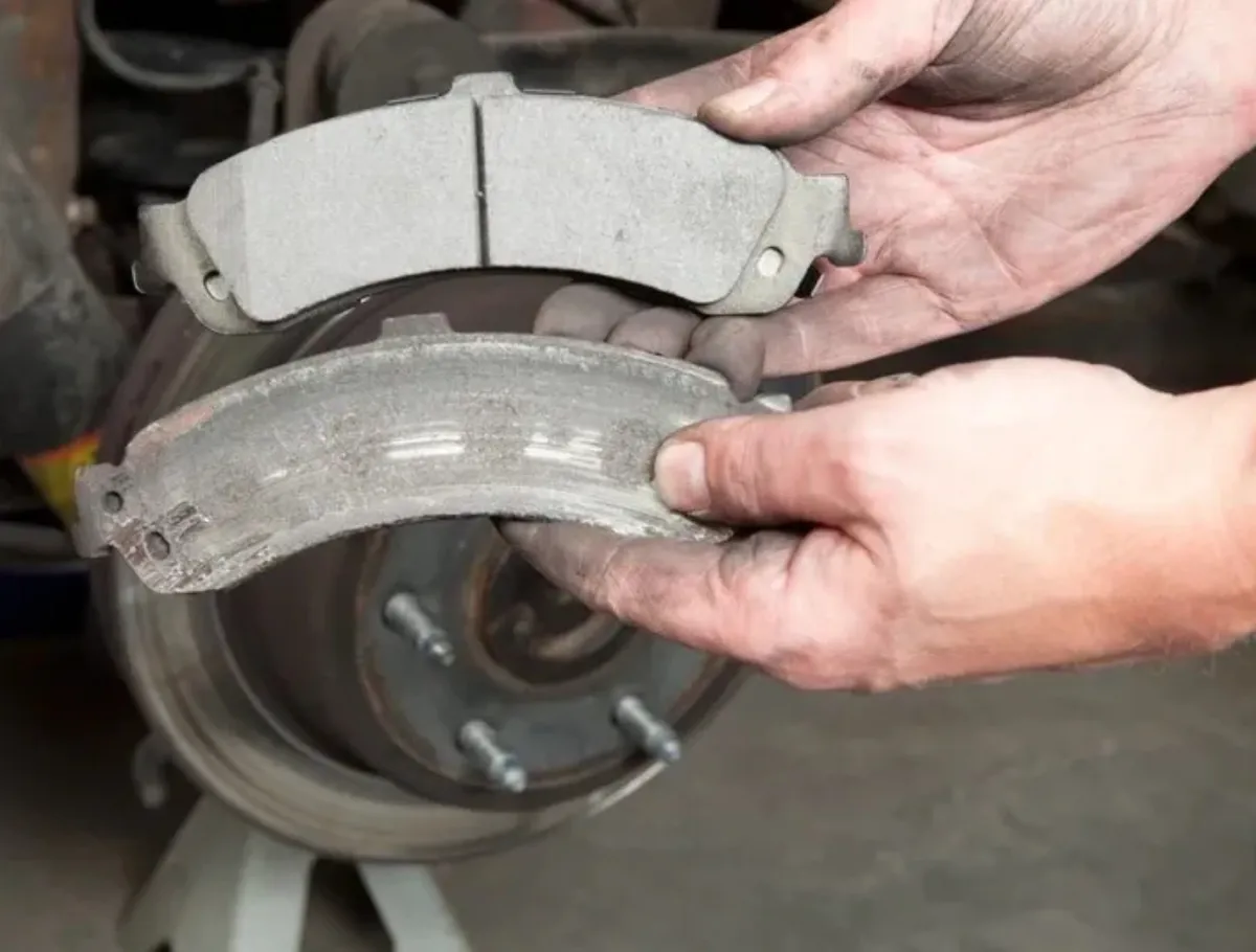 Hands holding a new brake pad above a worn brake pad near a car's brake rotor.
