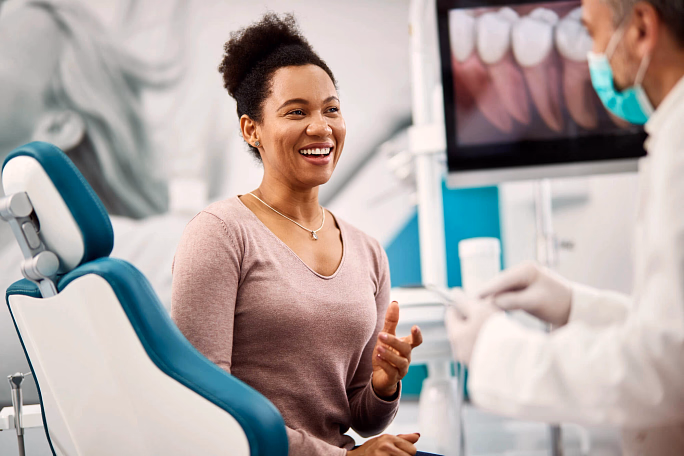 Smiling woman sitting in a dental chair talking to a masked dentist showing a tooth model.